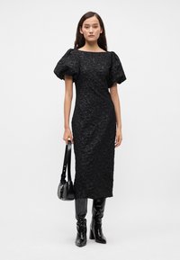 Crna obleka s puhastimi rokavi in teksturiranim cvetličnim vzorcem, oprijet silhuet in dolžina do kolen. Sestavljena z črnimi sijajnimi škornji do kolen in majhnim ročajem.