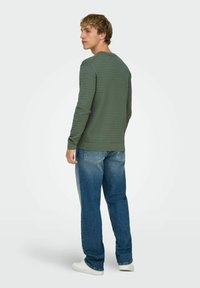 Chemise verte texturée à manches longues, col rond, associée à un jean en denim bleu clair et des chaussures blanches, vue de dos.