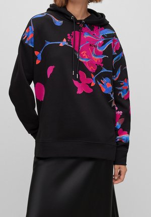 Zwarte hoodie met levendige roze en blauwe bloemmotieven, capuchon met trekkoord, gedragen met een zwarte rok, model getoond van nek tot heupen.