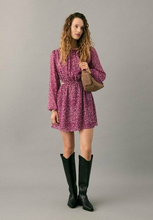 Purple patterned dress with long sleeves and a cinched waist, paired with black knee-high boots and a small brown bag.