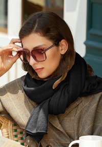 Sciarpa nera e strutturata drappeggiata attorno al collo, abbinata a occhiali da sole oversize color maroon e un cappotto a scacchi color sabbia. Tazza di caffè in primo piano.
