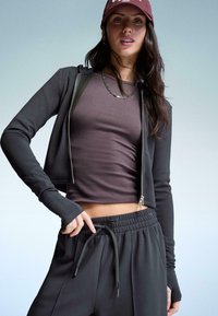 Fiatal nő bordó sapkában, lánc nyaklánccal, lila cropped toppal, fekete cipzáras dzsekivel és fekete húzós nadrággal, egy sima háttér előtt.