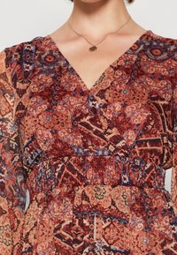 Langärmliges Kleid mit V-Ausschnitt in einem roten und blauen, filigranen Muster, das mit einem gerafften Taillen-Detail und leichtem, transparentem Stoff ausgestattet ist.