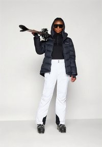 Musta toppatakki, valkoiset lasketteluhousut, mustat laskettelumonot, pidellen suksia, tyylikäs muotoilu, kontrastivärit, istuva siluetti, vetoketjut housuissa.