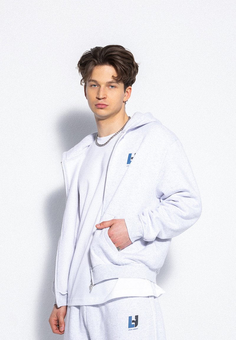 Jasnoszara, zapinana na zamek bluza z miękkiego materiału posiada kieszeń z przodu oraz logo na klatce piersiowej. Nosi ją w połączeniu z białą koszulką i dopasowanymi spodniami dresowymi.