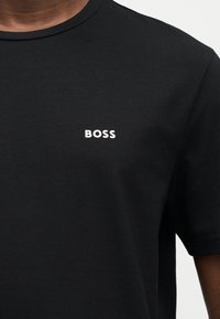 T-shirt noir à manches courtes avec le texte blanc "BOSS" centré sur la poitrine, porté par une personne dont le cou et l'épaule sont visibles.