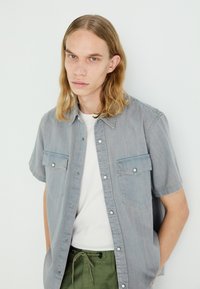 Camisa de denim azul claro de manga corta con dos bolsillos en el pecho y botones a presión, llevada sobre una camiseta blanca, combinada con pantalones verdes con cordones.