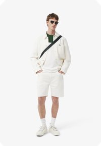 Chaqueta de mezclilla blanca, camiseta blanca, acento verde, pantalones cortos blancos y zapatillas blancas. Gafas de sol negras y correa cruzada negra.