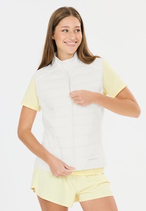 Jonge vrouw die glimlacht terwijl ze een witte mouwloze donsvest dichtritst over een lichtgele T-shirt en bijpassende korte broek, tegen een witte achtergrond.