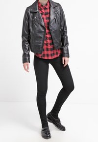 Giubbotto da moto in pelle nera, camicia a scacchi rossa e nera, jeans skinny neri e scarpe nere. Presenta cerniere sul giubbotto.