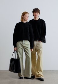 Δύο άτομα με μαύρα πουλόβερ και μπεζ παντελόνια φαρδιάς γραμμής στέκονται δίπλα δίπλα μπροστά σε ένα απλό ανοιχτό γκρίζο φόντο, το ένα κρατώντας μια μαύρη τσάντα ώμου.