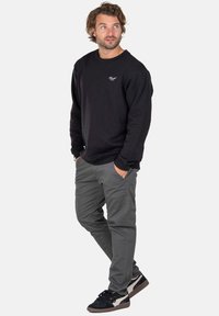 Mann mit Händen in den Taschen, der einen schwarzen Sweatshirt, graue Hose und schwarz-weiße Sneakers trägt und leicht nach rechts schaut.