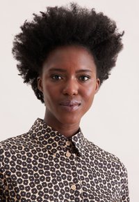 Giovane donna nera con capelli afro naturali, che indossa una camicia button-up a righe beige e nere, in posizione frontale con un'espressione neutra.