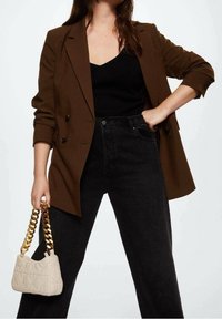 Femme portant un blazer marron, un top noir et un jean noir, tenant un sac à main beige matelassé avec une épaisse chaîne en or.