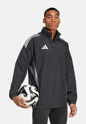 Zwarte sportjack met witte strepen, met rits aan de voorkant en elastische boorden, gedragen door een model dat een zwart-wit voetbal vasthoudt.