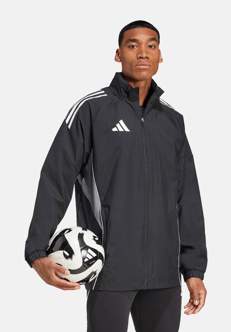Zwarte sportjack met witte strepen, met rits aan de voorkant en elastische boorden, gedragen door een model dat een zwart-wit voetbal vasthoudt.