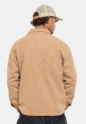 Homme portant une veste beige avec un logo embossé "Von Dutch California" dans le dos et une casquette de baseball en maille beige, vu de dos.
