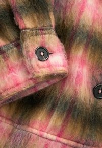 Gros plan d’un tissu en laine à carreaux rose, marron et crème avec un bouton noir cousu sur un poignet.