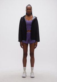 Femme debout, de face, portant une veste noire, un brassière de sport violette, un short, des chaussettes blanches et des baskets blanches sur un fond uni.