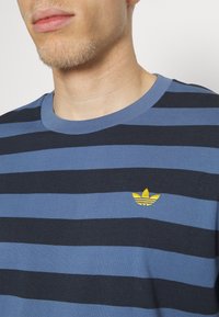 Man i en blå och svart randig skjorta med ett gult broderat märke på bröstet.