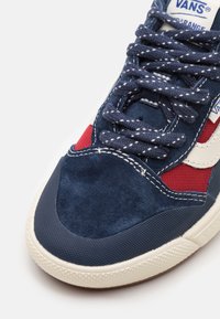 Ingrandimento di una sneaker Vans blu, rossa e bianca con suola testurizzata, lacci blu con punti bianchi e dettagli in tessuto scamosciato e mesh.