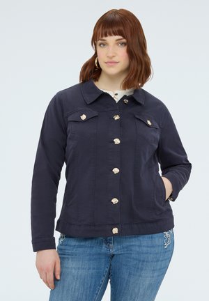 Frau mit roten Haaren, die eine marineblaue Jacke mit einzigartigen goldenen Knöpfen und blaue Jeans trägt, posiert mit einer Hand in der Tasche.