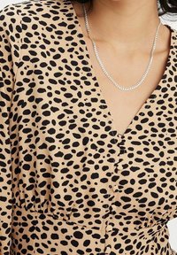 Beige jurk met zwart gespikkeld patroon, met een V-hals, korte mouwen en knoopdetails aan de voorkant. Zilveren kettinghanger zichtbaar.