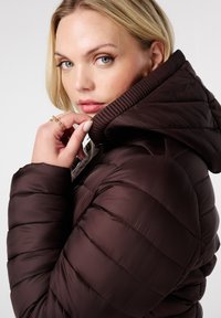 Blonde vrouw met lichte ogen, gekleed in een donkerbruine pufferjack, houdt de kraag bij haar kin en toont gouden ringen op haar vingers.