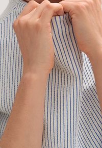 Vitt tyg med vertikala blå ränder, hålls av två händer. Texturen verkar mjuk och något skrynklig, med synliga händer som greppar materialet.