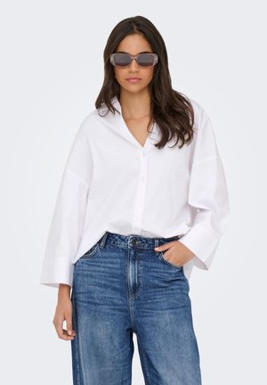Femme aux longs cheveux bruns portant une chemise blanche ample à boutons, un jean bleu et des lunettes de soleil rectangulaires foncées, une main dans la poche.