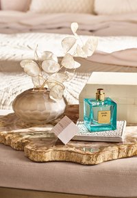 Bouteille de parfum turquoise transparente avec des accents dorés, sous-plat en céramique texturée, vase en verre gelé couleur noisette, et feuilles séchées sur un plateau en bois.