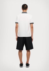 Biely polo tričko s čierne vzorovaným golierom a manžetami, v kombinácii s čiernymi šortkami. Obuv má čierne akcenty a jednoduchý dizajn.