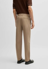 Beżowe spodnie na miarę wykonane z gładkiego materiału, z prostymi nogawkami, szlufkami na pasek i dwoma tylnymi kieszeniami. Widoczne czarne obuwie.