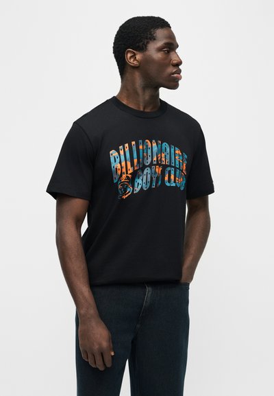 Άνδρας με μαύρο μπλουζάκι που φέρει πολύχρωμο κείμενο και γραφικό "Billionaire Boys Club", στέκεται με το ένα χέρι στην τσέπη, κοιτάζοντας δεξιά.