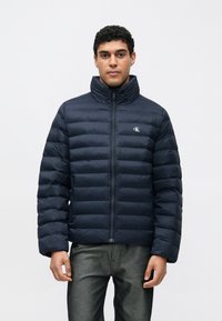 Geacă puffer bleumarin cu guler înalt, design matlasat, fermoar frontal și logo discret pe piept. Textură netedă și ușoară.