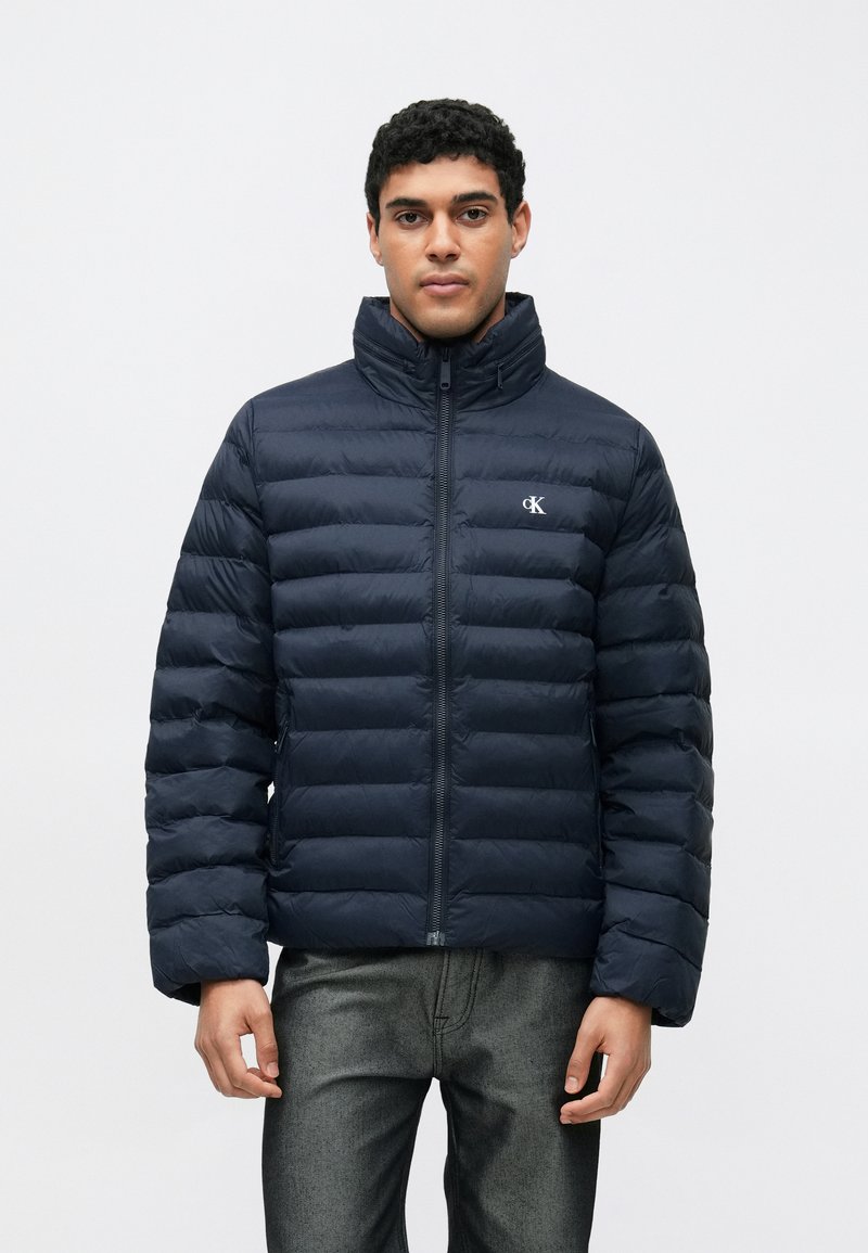 Geacă puffer bleumarin cu guler înalt, design matlasat, fermoar frontal și logo discret pe piept. Textură netedă și ușoară.