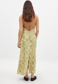 Robe maxi en tissu jaune pâle avec des motifs de roses en 3D. Dos ouvert avec des fines bretelles, présentant une fermeture à nouer et une fente sur le côté. Chaussures plates noires.