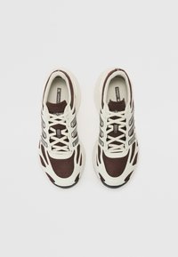 Baskets de sport marron et crème avec une tige en maille et des panneaux texturés, présentant des rayures noires et une semelle arrondie. Les lacets sont bien serrés.