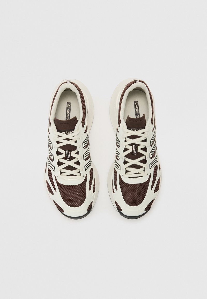 Braune und cremefarbene Sportschuhe mit einem oberen Teil aus Mesh und strukturierten Paneelen, versehen mit schwarzen Streifen und einer abgerundeten Sohle. Die Schnürsenkel sind fest gebunden.