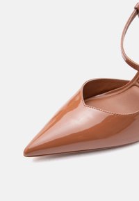 Chaussure pour femme brillante couleur caramel, à bout pointu, avec bride à la cheville et finition lisse, vue en gros plan sur fond blanc.