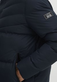 Marineblå puffjakke med quiltet design, sidelommer med lynlås og stofpatch-logo på venstre ærme. Blød tekstur, let udseende.