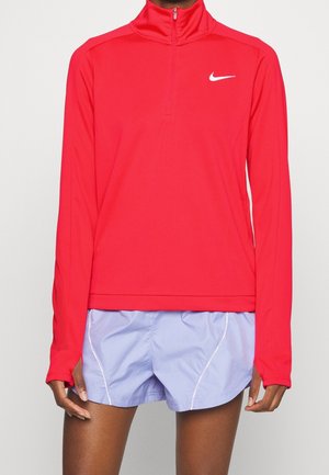 Personne portant un haut Nike rouge à manches longues avec fermeture éclair sur le quart et un short de course violet clair sur un fond uni.