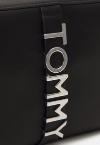 Borsa in pelle nera con una tracolla metallica con il logo "TOMMY", con una texture liscia e un design elegante. Accenti di hardware argento visibili.