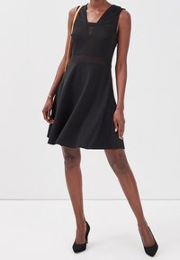 Personne mince en robe noire sans manches arrivant aux genoux avec des panneaux transparents, portant des talons noirs et portant un sac à bandoulière avec des sangles jaunes.