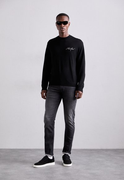 Suéter negro de manga larga con bordado blanco, jeans grises y zapatillas negras. El modelo lleva gafas de sol negras y está de pie contra un fondo neutral.