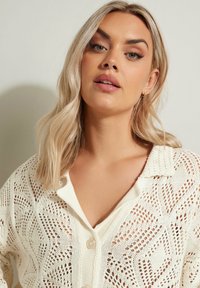 Stickad krämfärgad kofta med öppet mönster, med en kragkrage och knappar ner framtill. Texturerat design genom hela plagget.