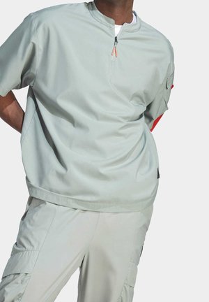 Mężczyzna w jasnoszarej koszulce z krótkim rękawem zapinanej na zamek przy szyi oraz dopasowanych spodniach cargo, stoi z rękami za plecami na jednolitym tle.