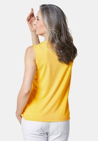 Top giallo senza maniche con una consistenza liscia, scollatura rotonda e vestibilità comoda, abbinato a jeans bianchi. Nessun motivo o dettaglio visibile.
