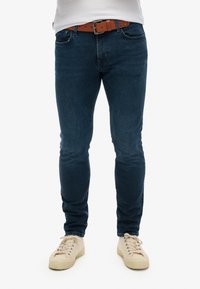 Jeans en denim bleu avec une coupe slim, accompagnés d'une ceinture marron. Chaussant des baskets beiges avec une surface texturée et des lacets clairs.