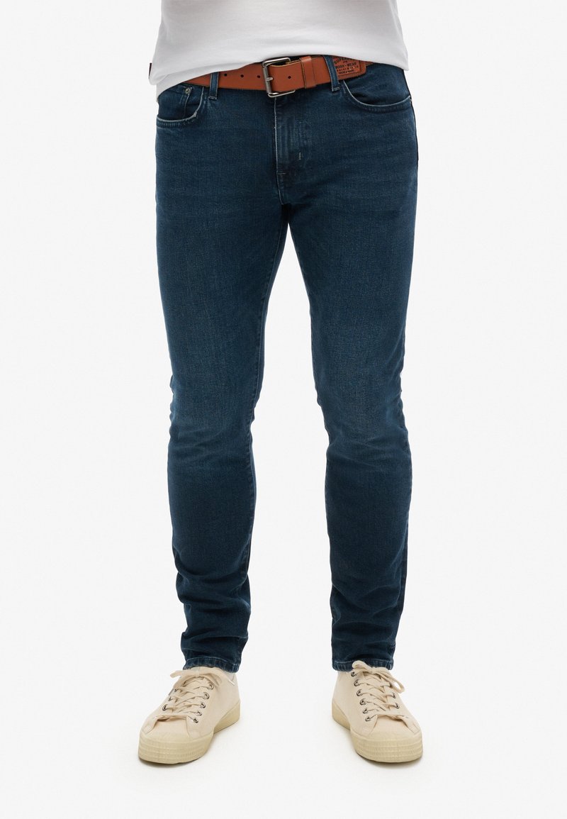 Jeans en denim bleu avec une coupe slim, accompagnés d'une ceinture marron. Chaussant des baskets beiges avec une surface texturée et des lacets clairs.